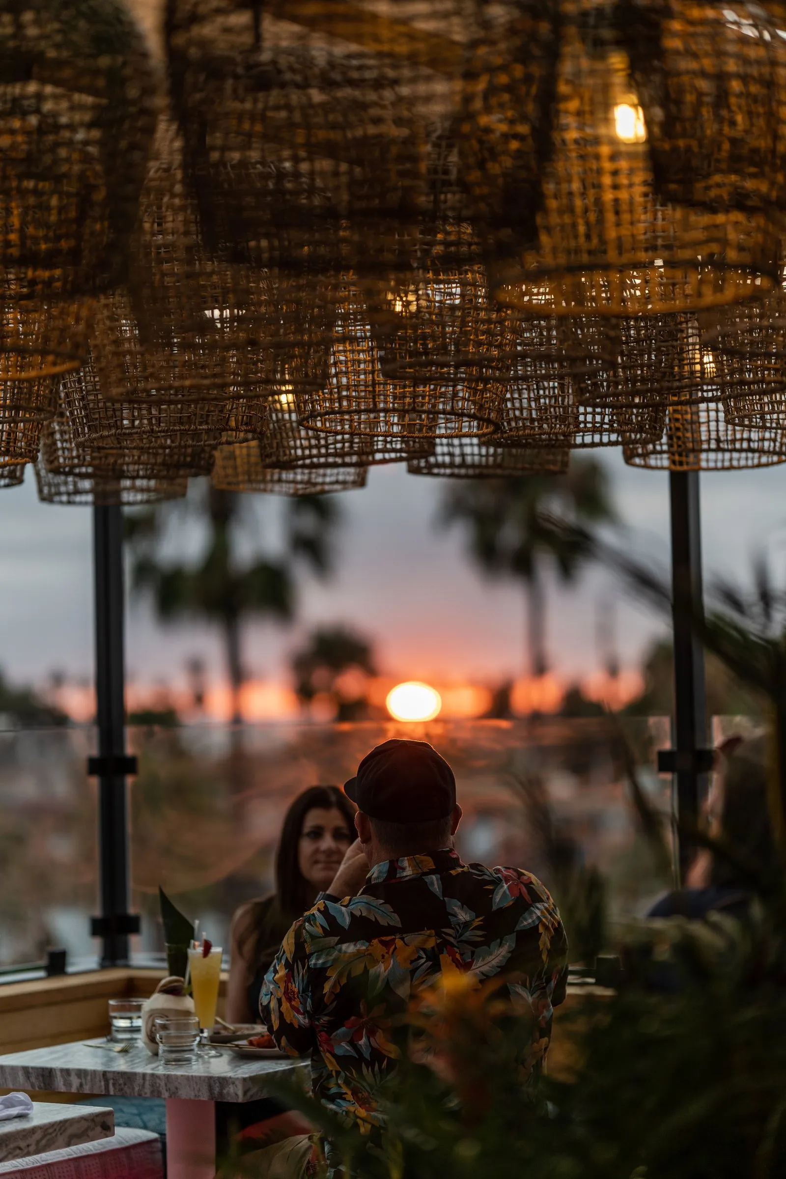 Sunset viewed from Coco Cabana rooftop through glass railing, palms silhouetted against orange sky